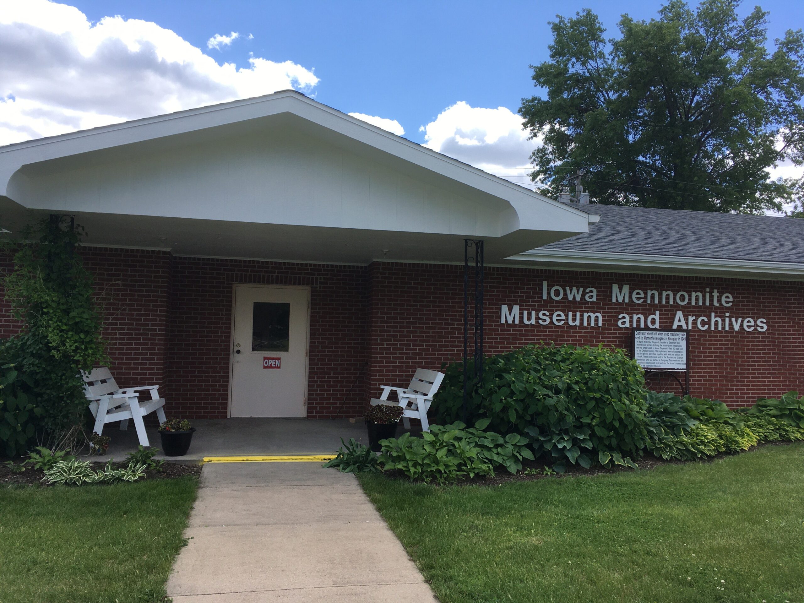 Front view of the Iowa Mennonite Museum and Archives. A low brick building with a white door and a gently-sloped shingled roof. "Iowa Mennonite Museum and Archives" is on the brick in white sans-serif letters.