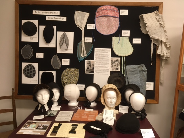 A display of Amish and Mennonite Head coverings. Numerous bonnets and other head coverings hang on a black cloth pinboard. In front of the pinboard, seven styrofoam heads model five black bonnets, a cream colored bonnet, and a white bonnet. Labels and photo illustrations are also featured.